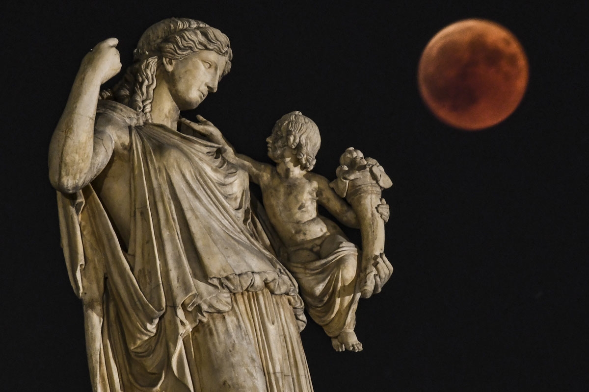 View of the full moon during the "blood moon" next to the ancient Greek goddess Irine holding a child symblolising peace and justice in central Athens on July 27, 2018. The period of total eclipse, when earth's shadow is directly across the moon and it is