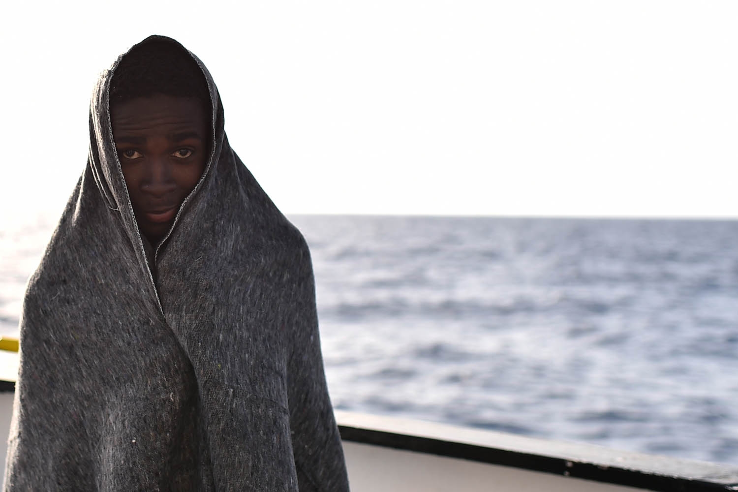A young man stands wrapped in a blanket after a rescue operation of refugees and migrants at sea of the rescue ship Aquarius, on May 24, 2016 in front of the Libyan coast