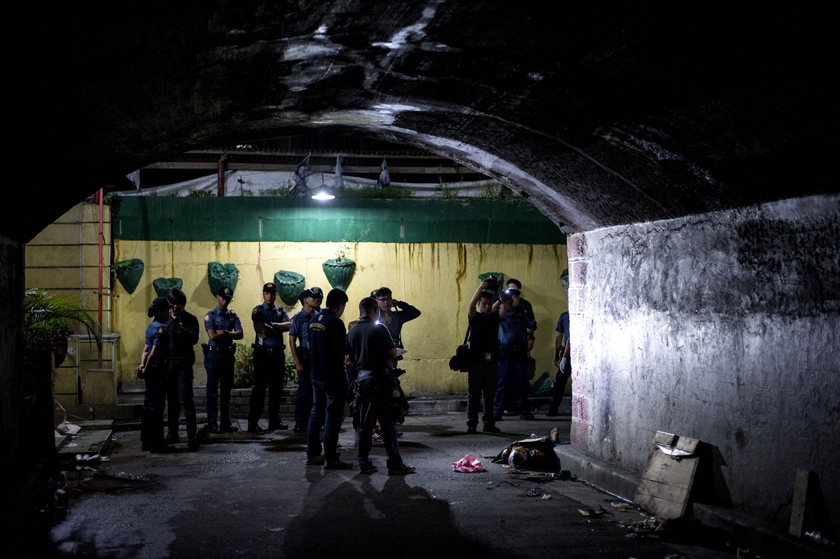 Police officers investigate the dead body of an alleged drug dealer (R), his face covered with packing tape and a placard reading I'm a pusher, on a street in Manila