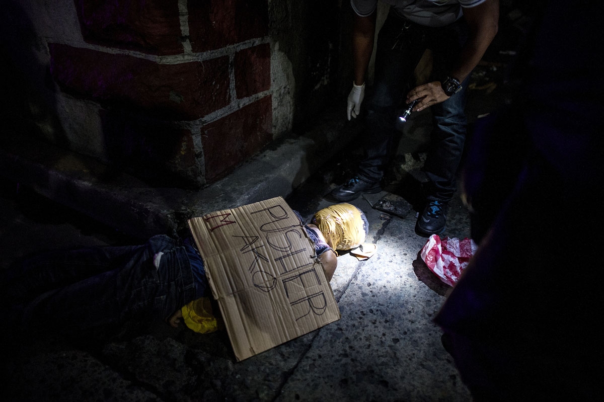 This photo taken on July 8, 2016 shows police officers investigating the body of an alleged drug dealer with his face covered with packing tape, with a cardboard sign on him reading I'm a pusher, at a street in Manila.