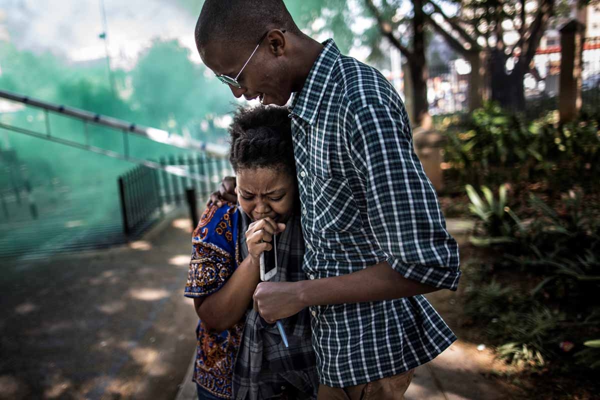 A wounded student is comforted by a fellow student as green smoke screen used by the South African Anti Riot Unit is seen in the background at a protest in Johannesburg on September 21, 2016. A wounded student is comforted by a fellow student as green smoke screen used by the South African Anti Riot Unit is seen in the background at a protest in Johannesburg on September 21, 2016.