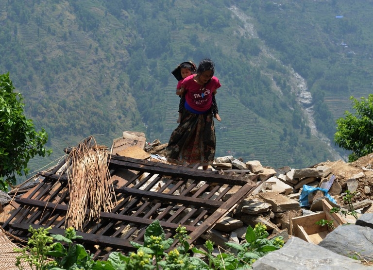 A Nepalese woman carries a child on her back past damaged homes in a village northwest of Kathmandu on May 11, 2015 A Nepalese woman carries a child on her back past damaged homes in a village northwest of Kathmandu on May 11, 2015